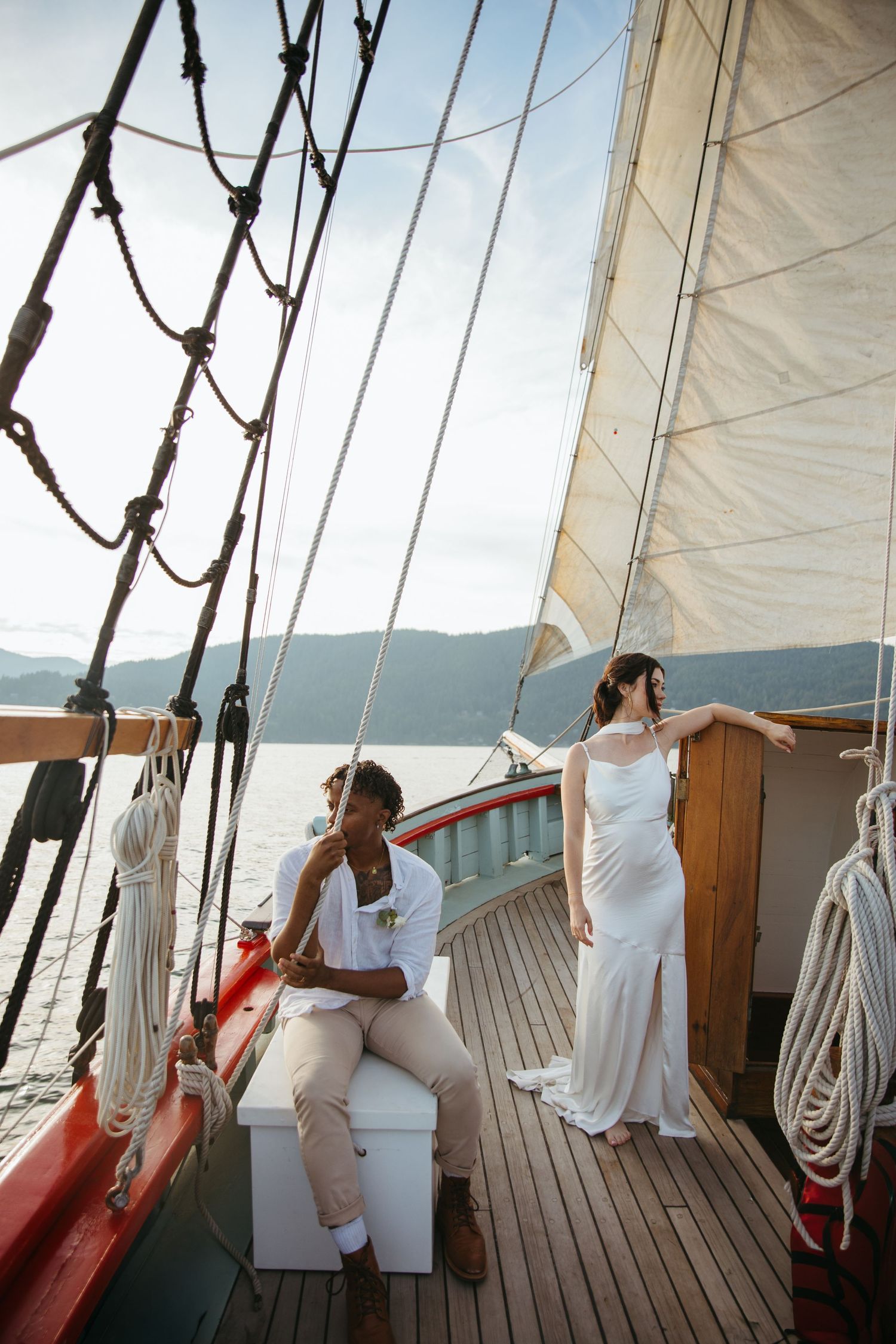 Bride and groom on a classic sailboat deck with white sails and mountain views on their wedding day.