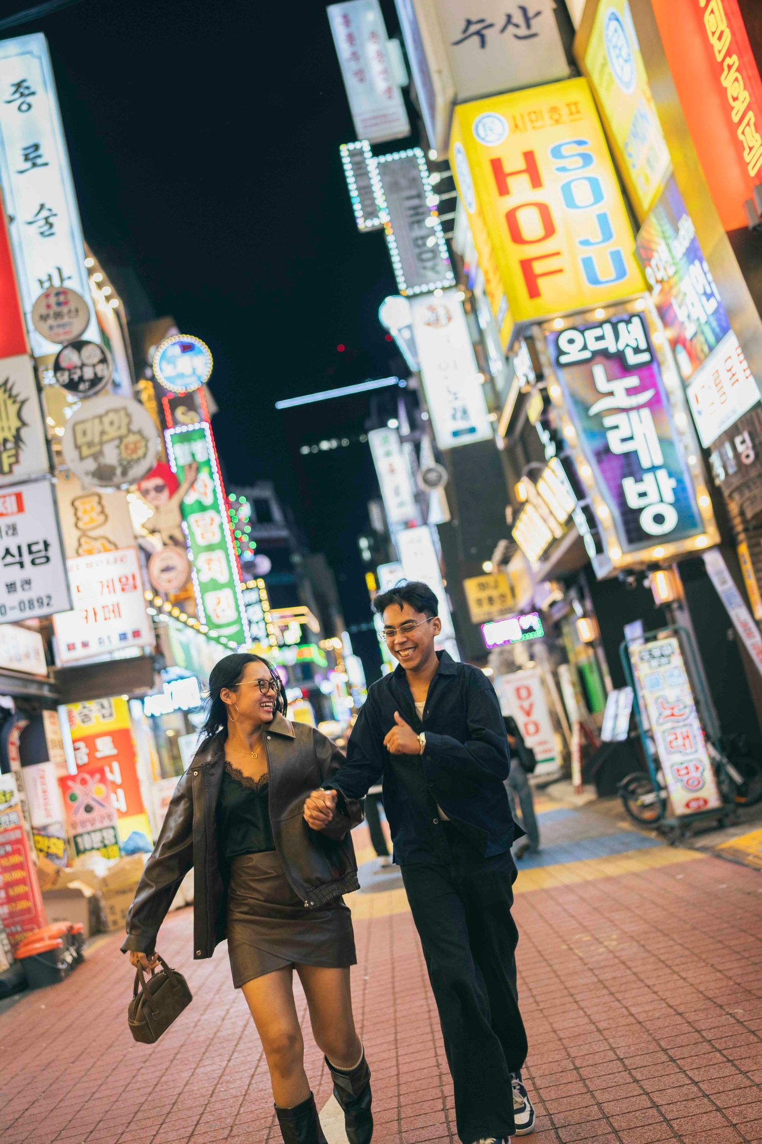 Couple laughing and walking together at night on a vibrant Seoul street lined with colorful Korean neon signs.