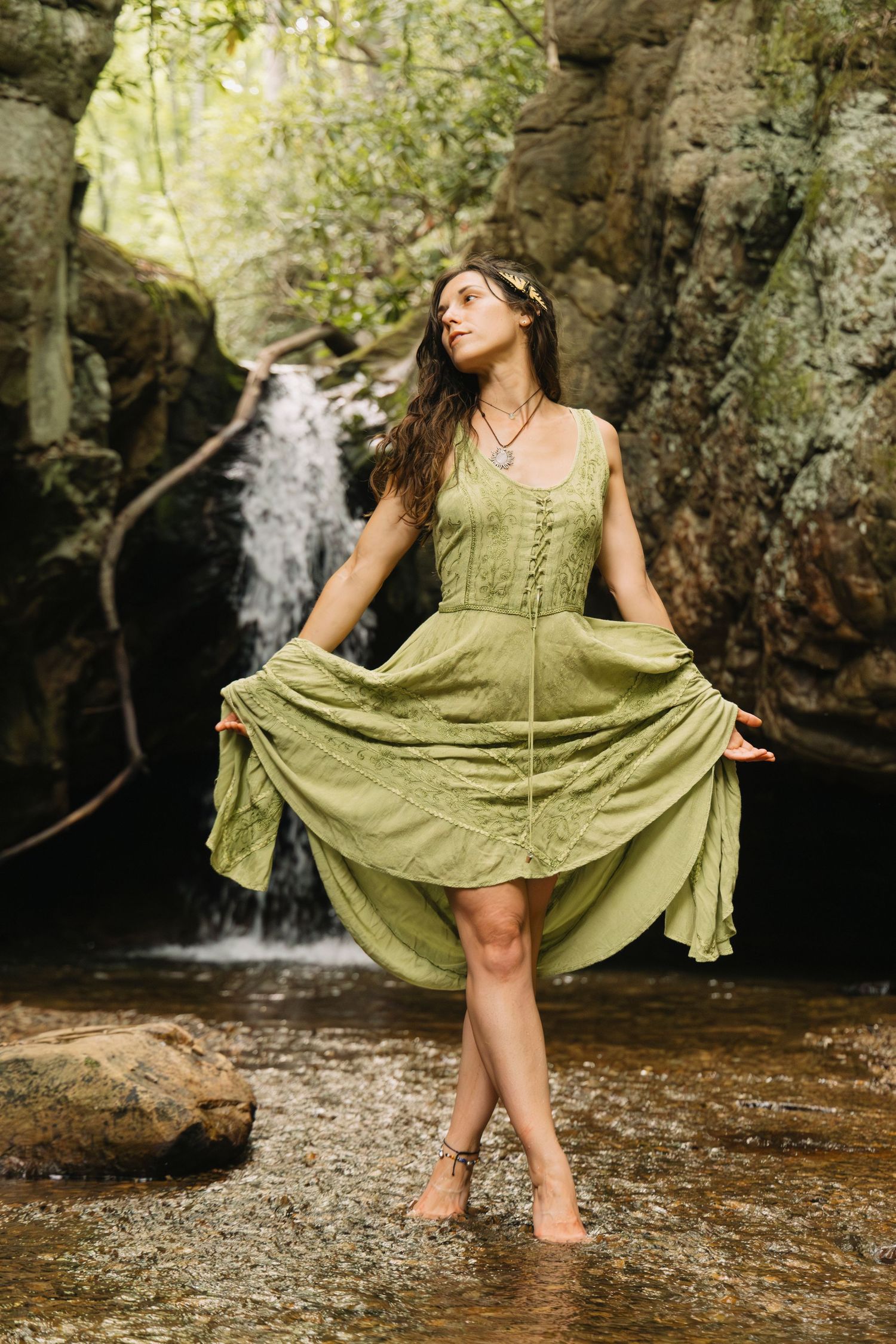 Woman in green dress standing in forest stream near waterfall, wearing floral crown, lifting skirt gracefully.