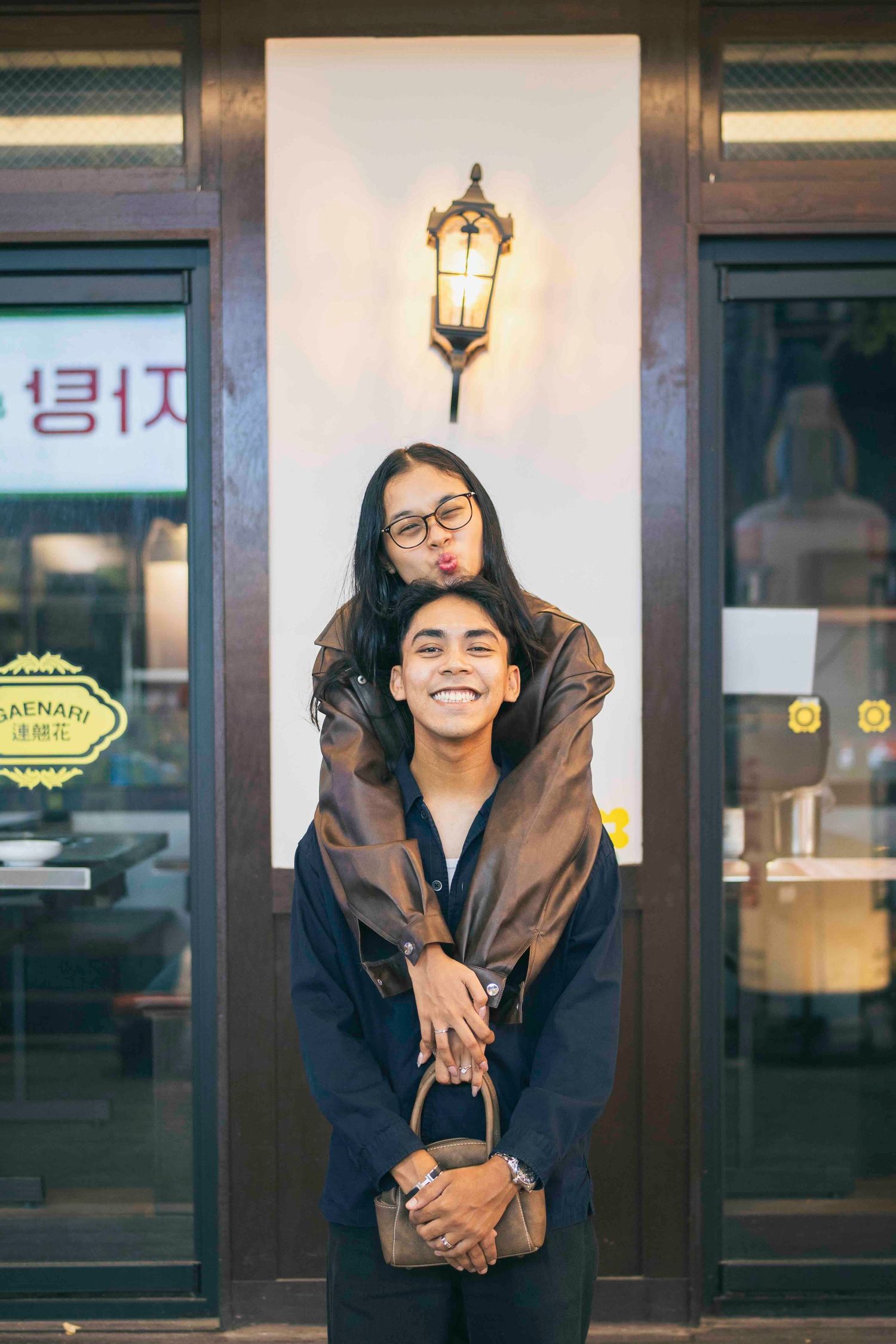 A smiling couple poses playfully outside a Korean shop, woman resting chin on man's head wearing brown leather jacket.