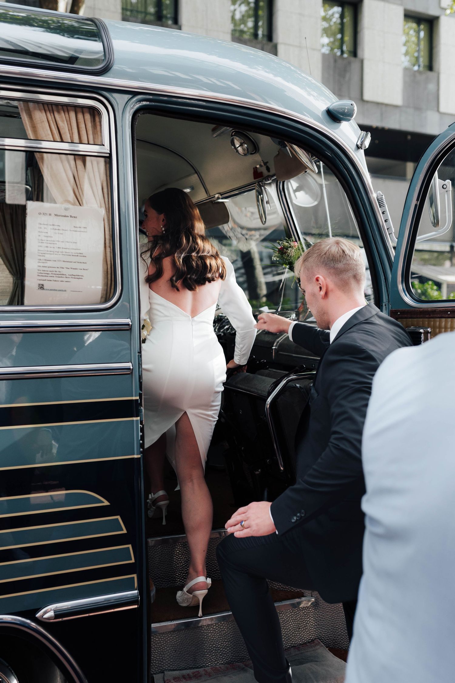 Bride in white dress boarding a vintage dark green bus assisted by groom in black suit on wedding day.
