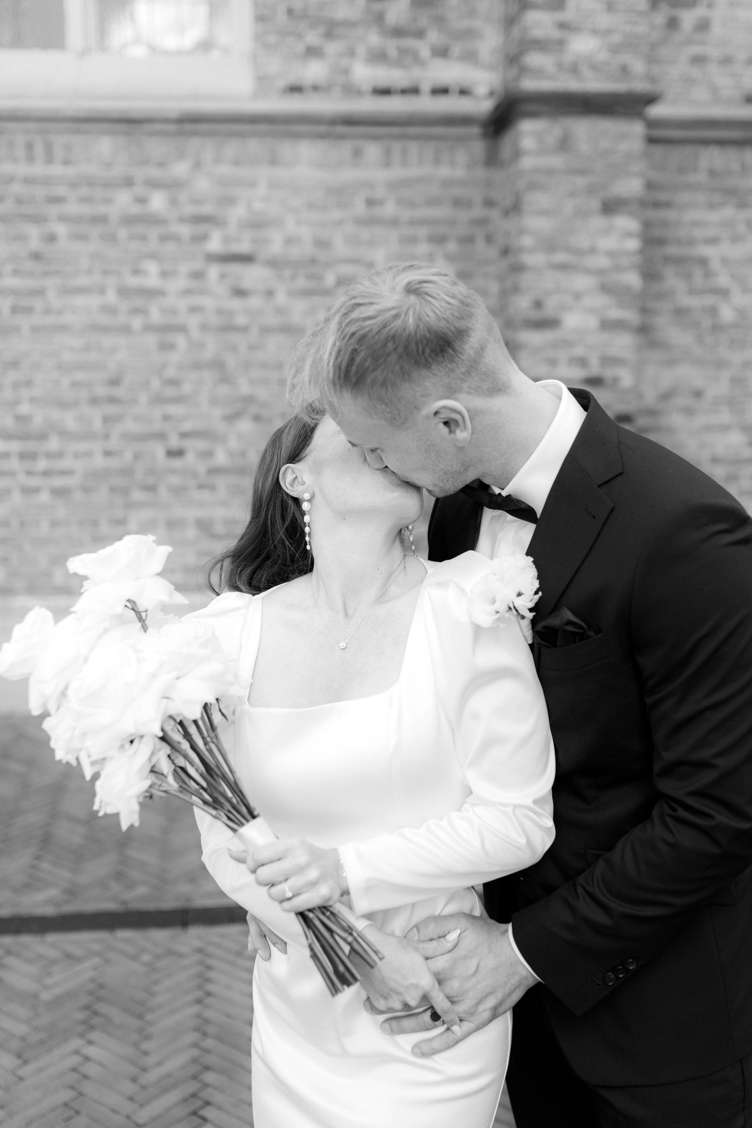 Black and white photo of a bride and groom kissing, bride holding white floral bouquet, brick wall background.