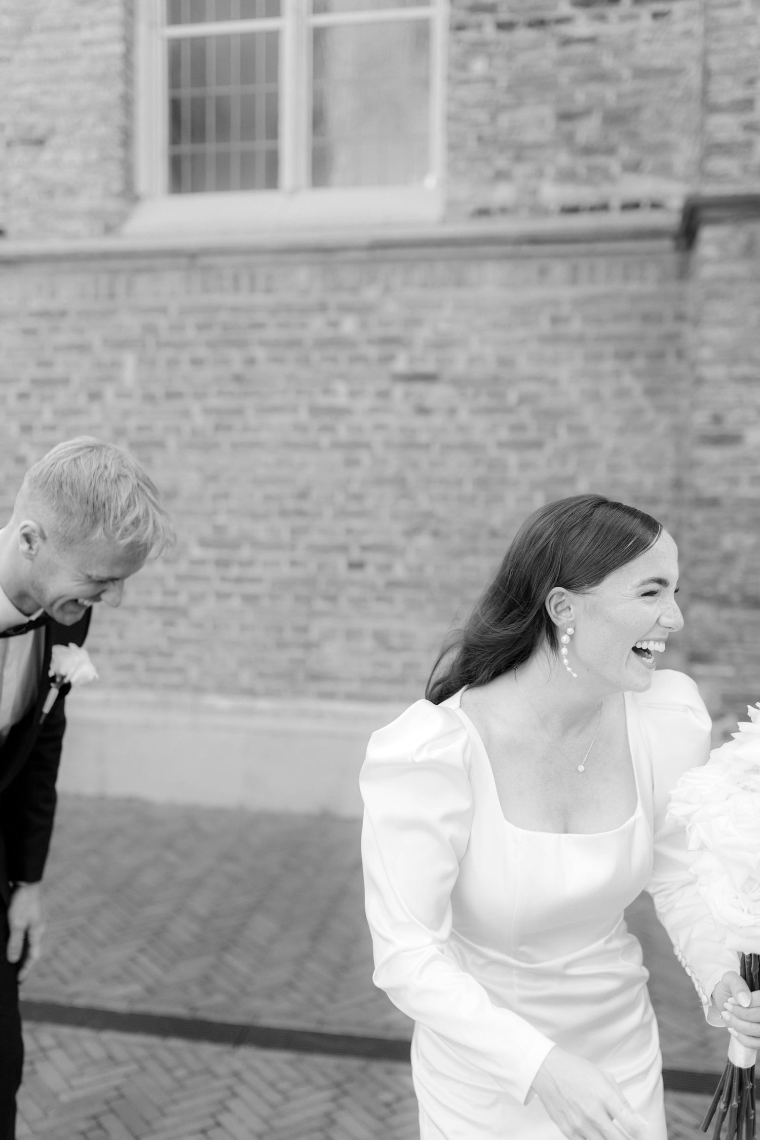 Black and white photo of a laughing bride in a white gown holding flowers beside a groom in a tuxedo near a brick wall.