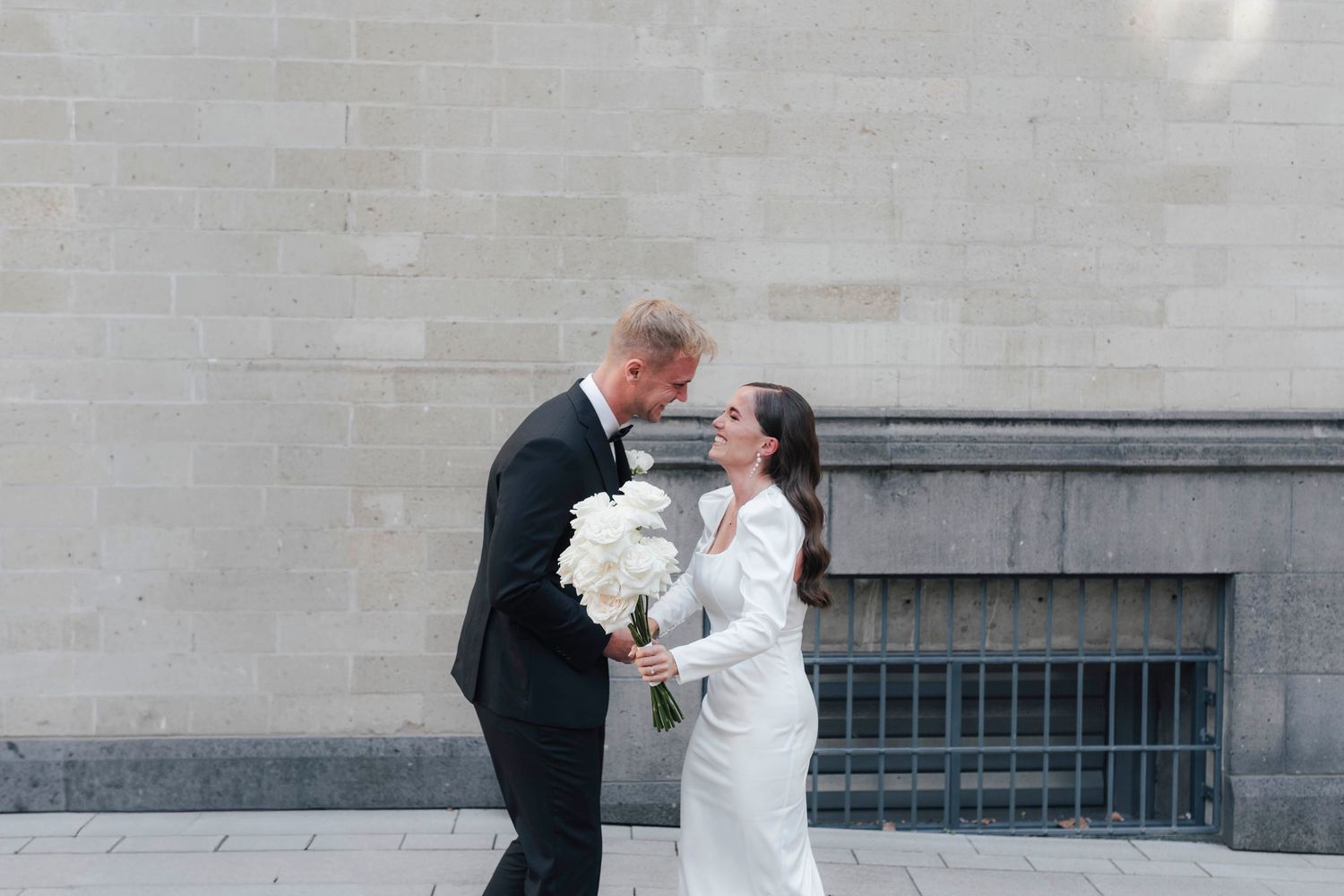 Bride and groom share a romantic moment outside, holding white peonies bouquet against a stone wall backdrop.