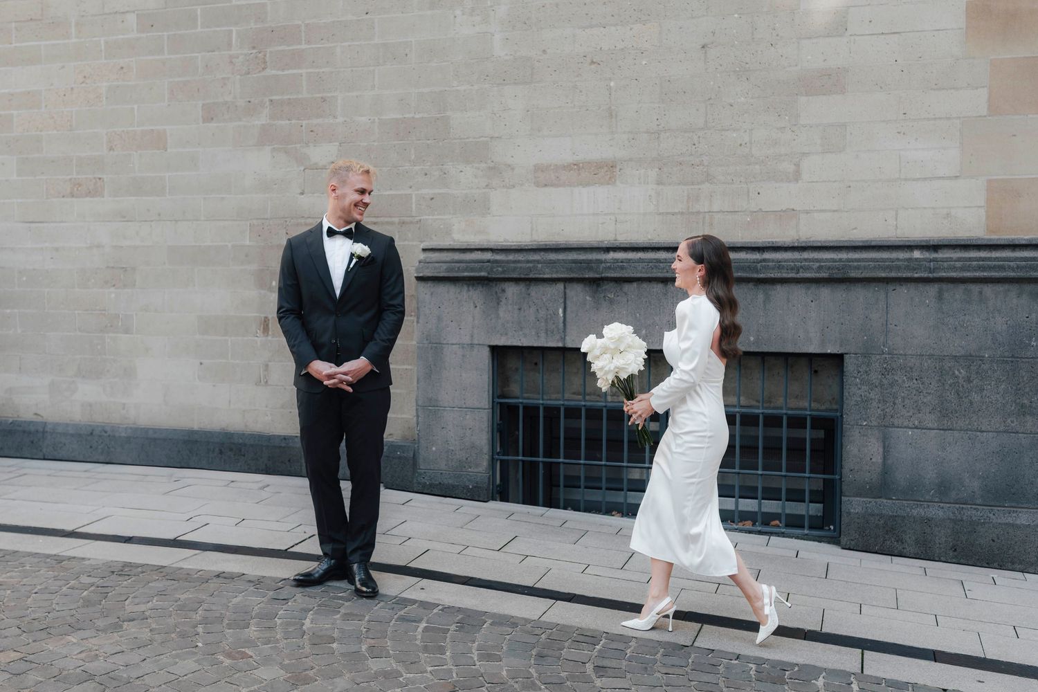 Bride in white dress carrying bouquet walks toward groom in black tuxedo against stone wall backdrop.