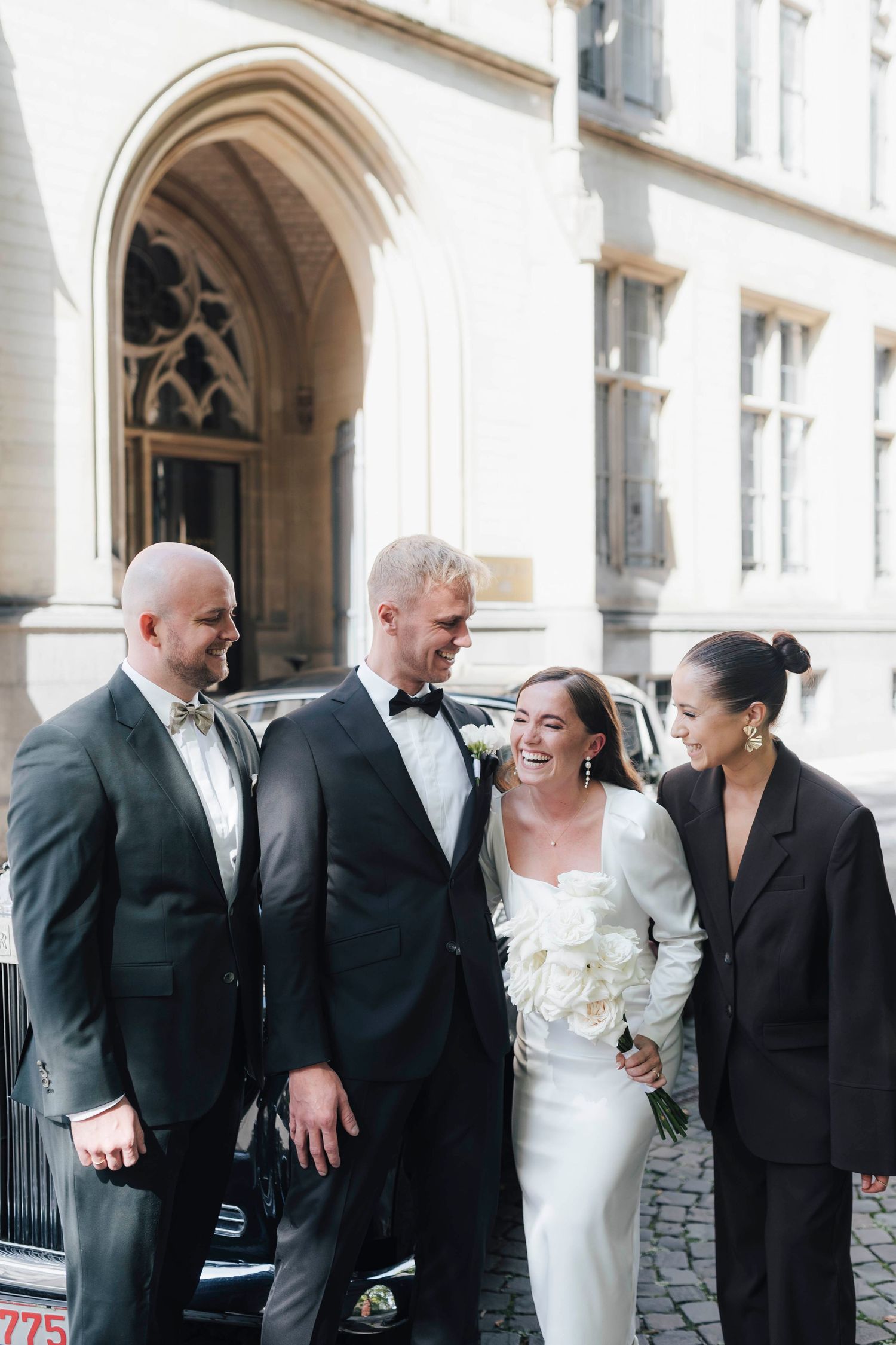 Bride in white gown with bouquet stands with groom in tuxedo and two guests outside elegant building.