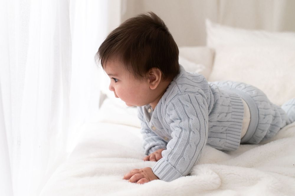 A cute baby in a blue quilted outfit crawling on a white bed near a bright window.