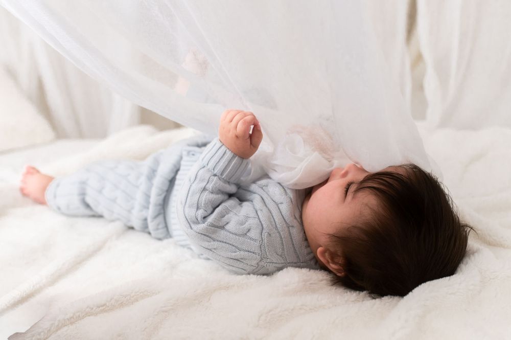 Peaceful baby in gray knit outfit lying on white bedding under a sheer canopy.