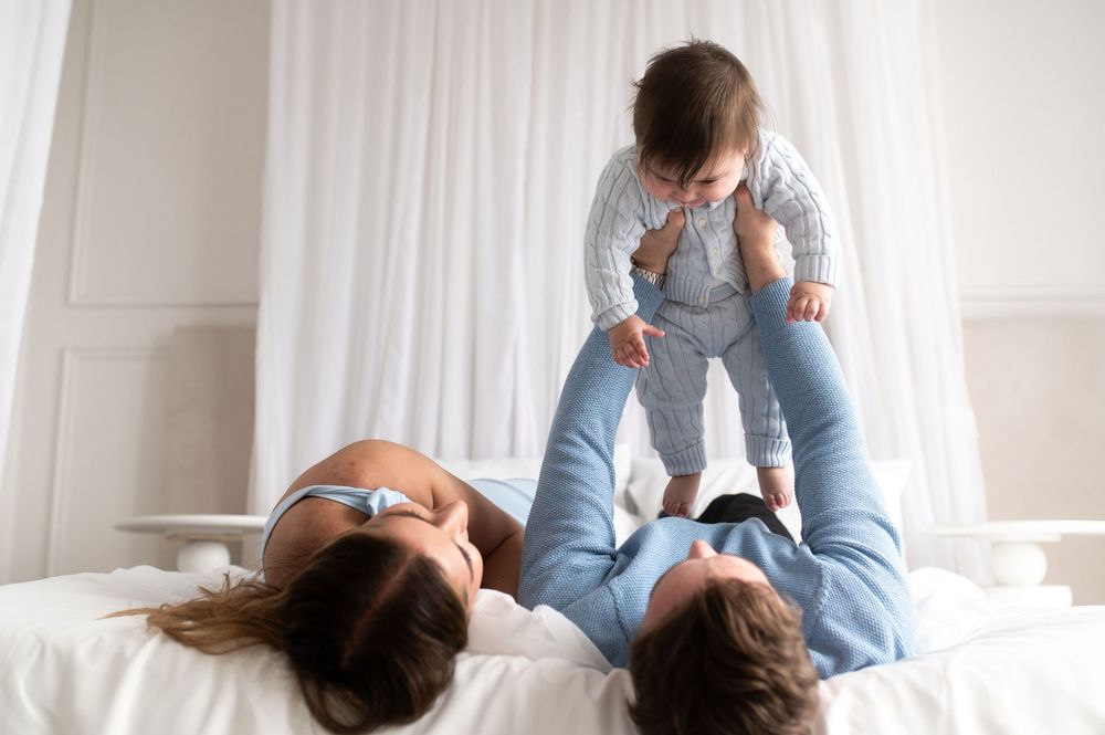 Parents lying on bed playfully lifting their baby in the air during a joyful family moment.