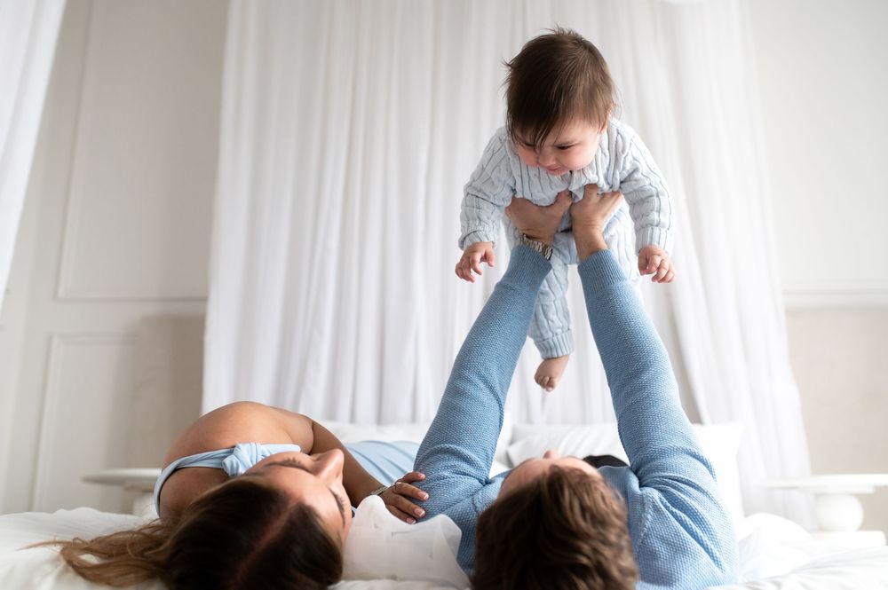 Mother lying on bed playfully lifting happy baby in the air during joyful bonding playtime at home.
