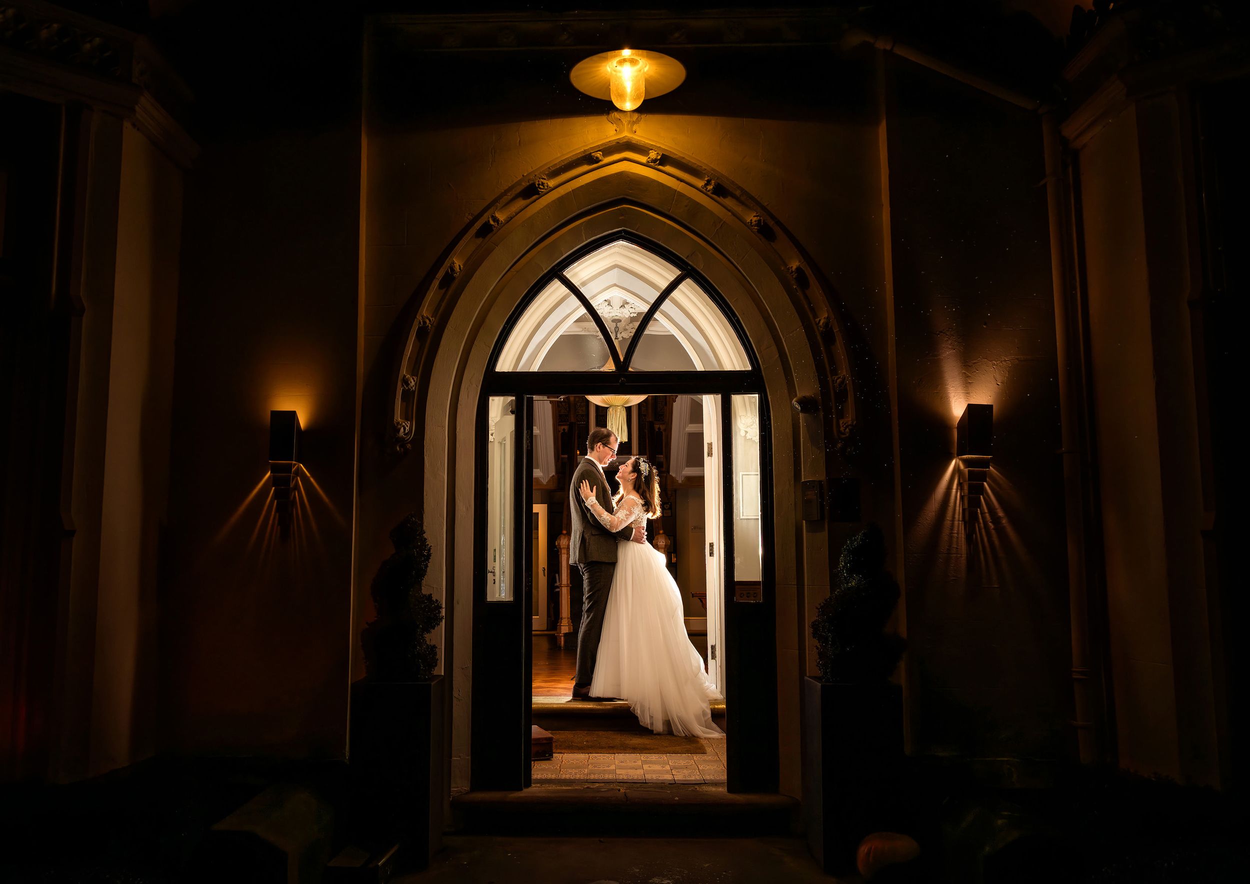 Bride and groom share a romantic kiss framed by a Gothic arch doorway in warm evening light.