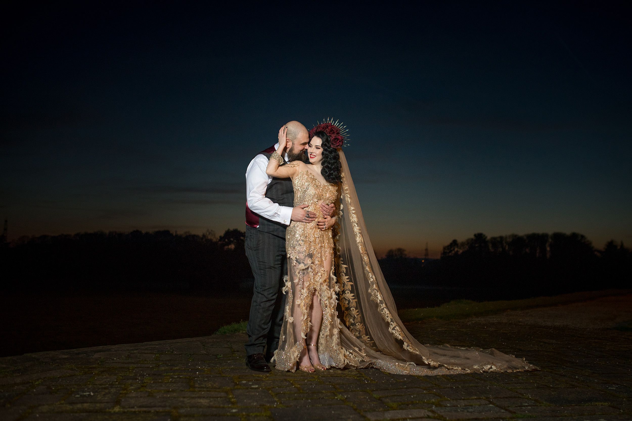 Couple in wedding attire share a romantic kiss at sunset, bride wearing gold embroidered gown with long veil.