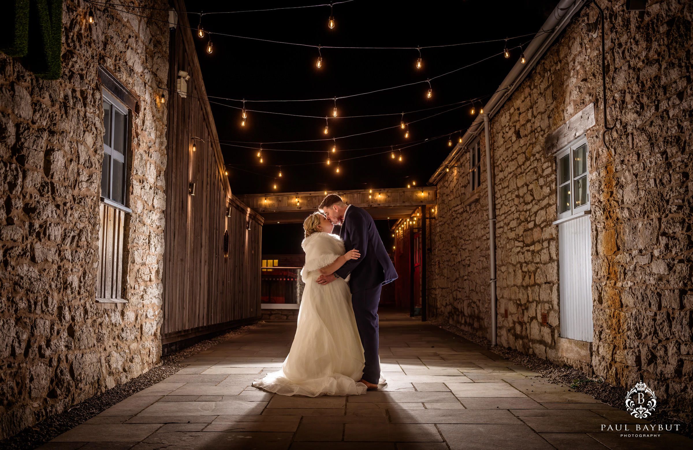 Bride and groom share a romantic kiss in a stone alley adorned with warm string lights at night.