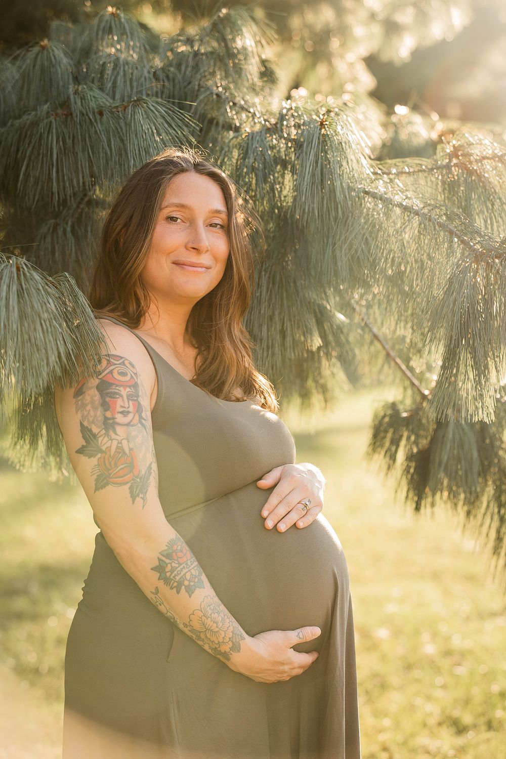 Expectant mother in gray dress poses in golden sunlight under pine branches.