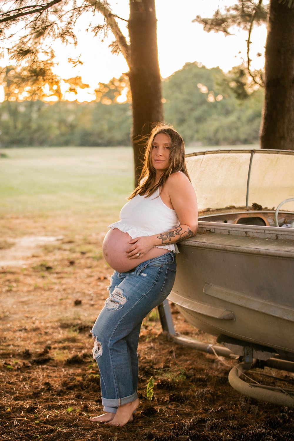 Pregnant woman in casual attire poses beside vintage car during sunset.