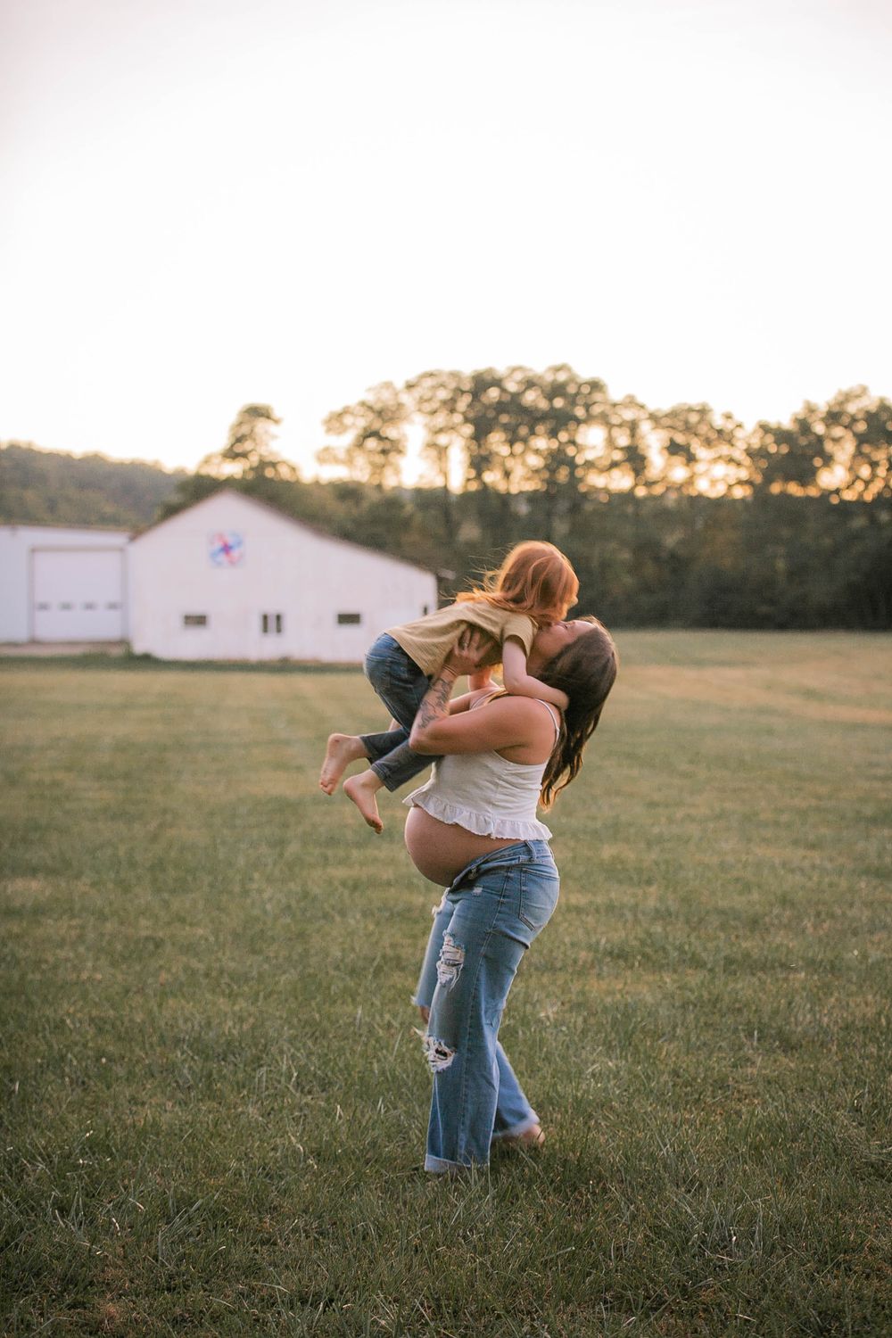 Couple embracing in field at sunset with white barn in background.