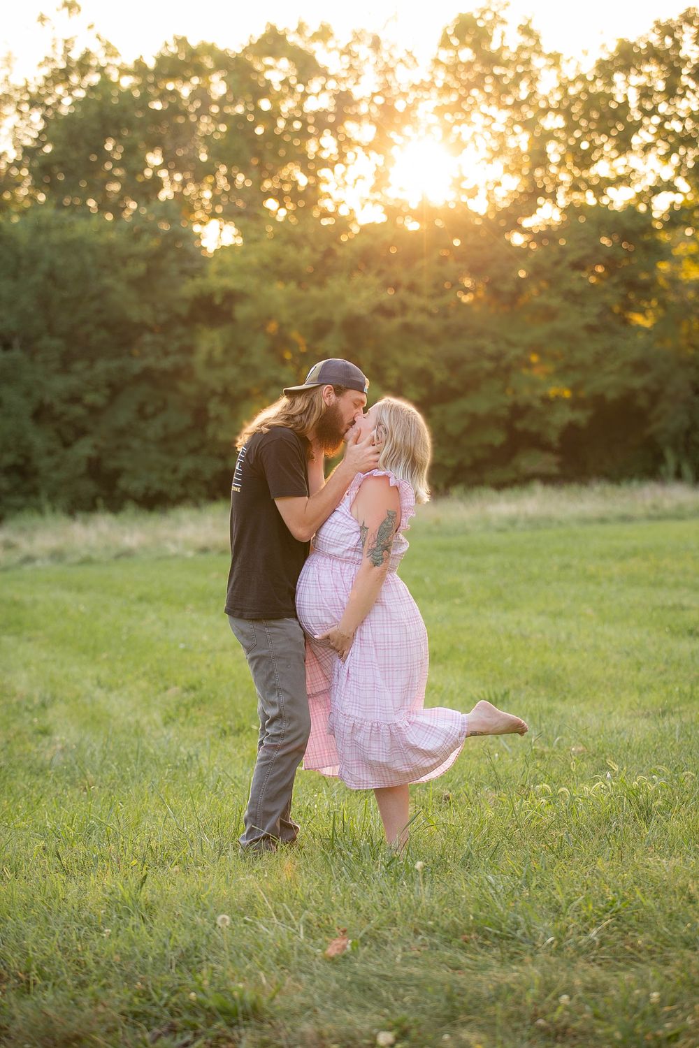 A romantic couple embraces in a grassy field during golden hour sunset with trees in the background.