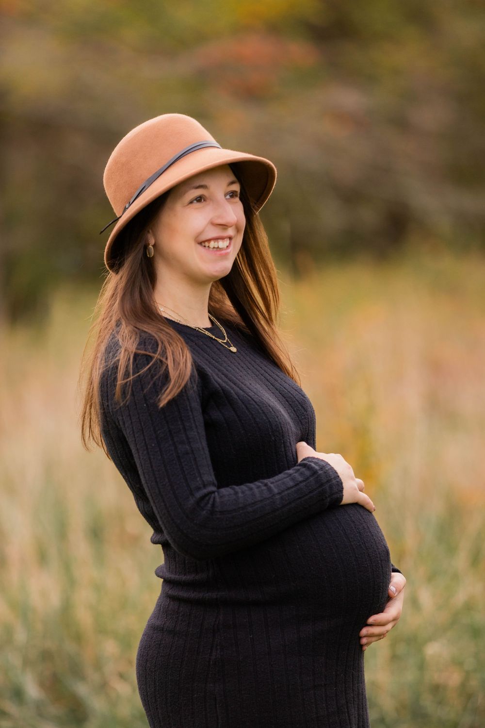 A person wearing a black sweater and brown hat poses in an autumn field during a maternity photoshoot.