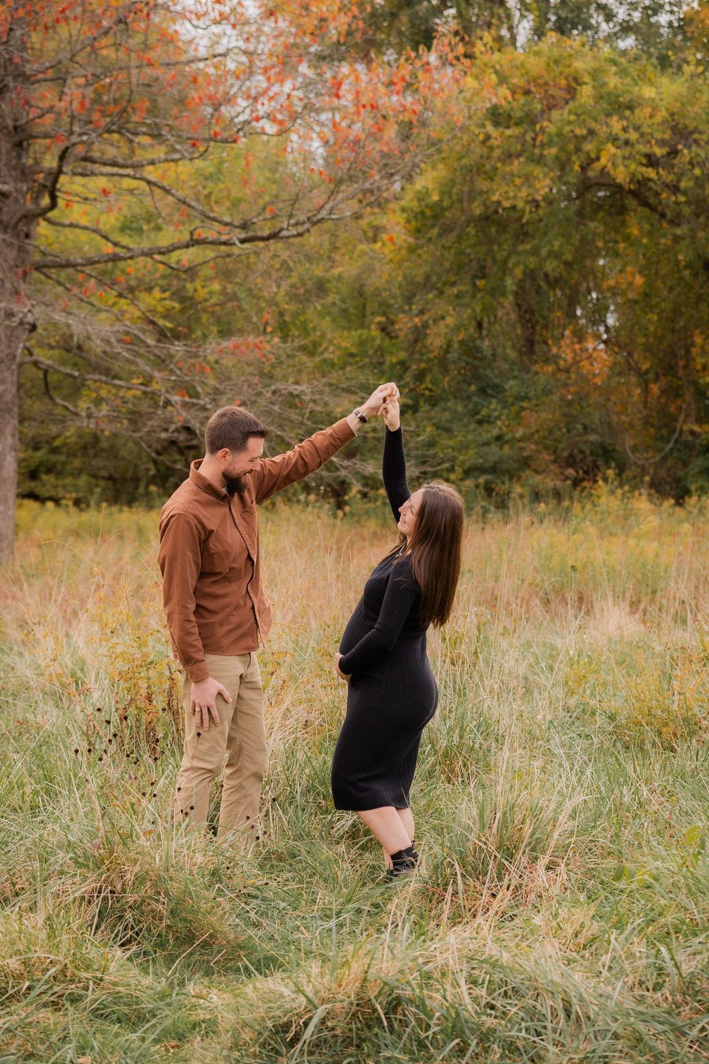 A couple dances playfully in a fall meadow surrounded by autumn trees with golden and orange foliage.