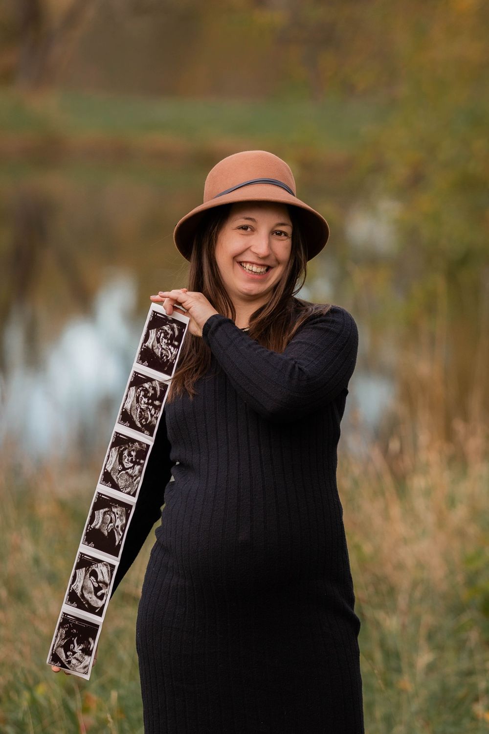 A pregnant person in a black sweater holds ultrasound photos while posing outdoors in autumn by a lake.