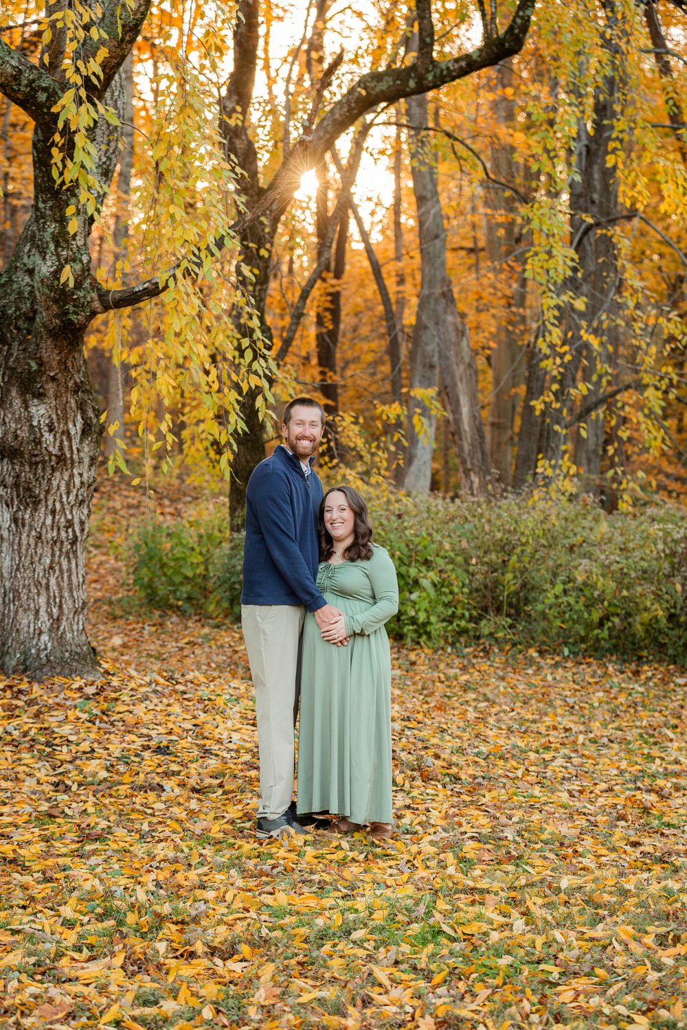 A couple stands together in a scenic autumn forest setting with golden leaves scattered on the ground during sunset.