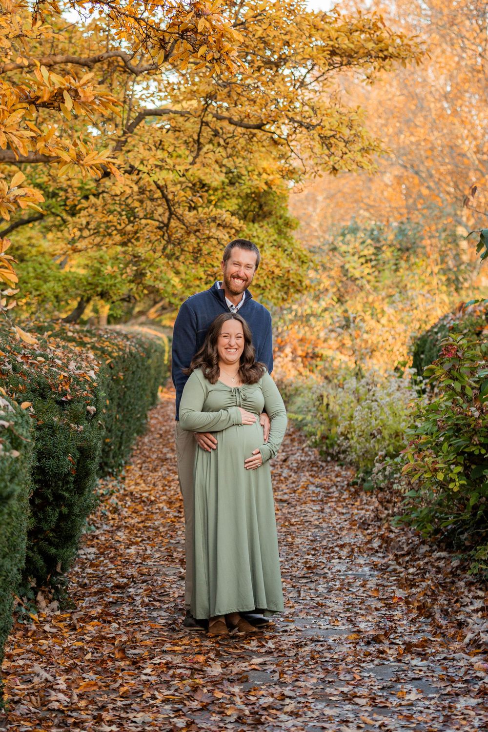 A couple in autumn woodland setting poses for maternity photos surrounded by golden fall foliage and scattered leaves.