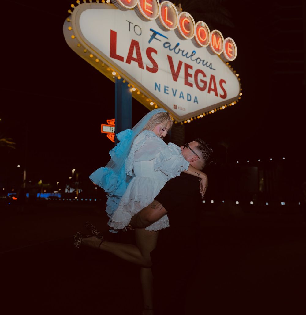 Couple celebrating at the iconic Welcome to Fabulous Las Vegas Nevada sign at night, man lifting woman in white dress.