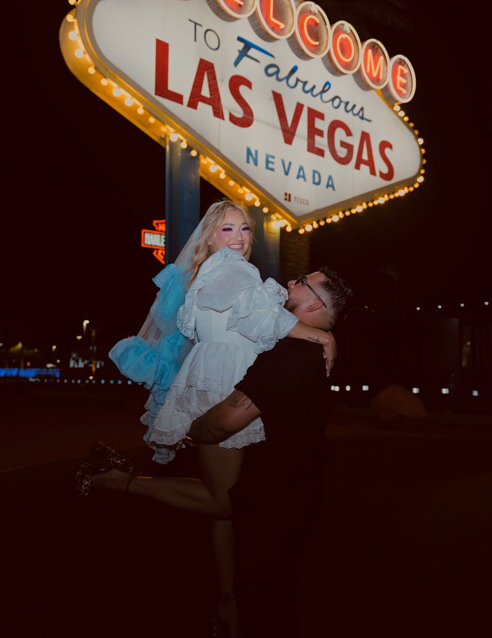 Joyful couple posing at the Las Vegas sign at night, man carrying woman in white dress and blue veil.
