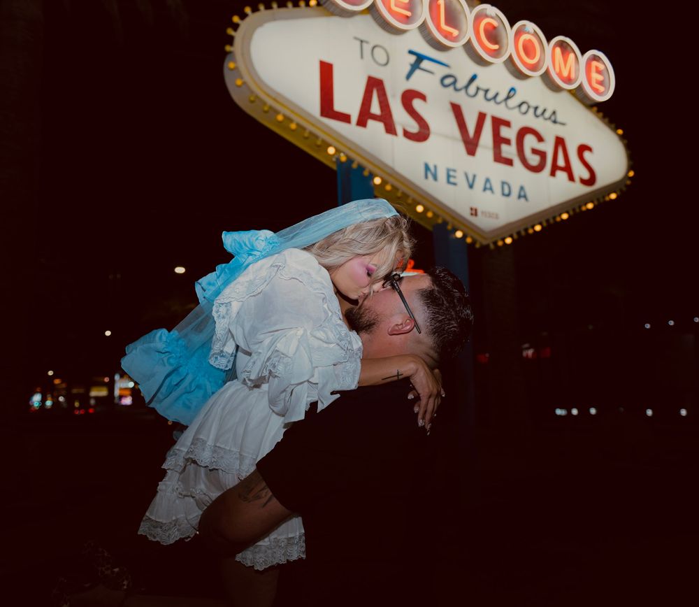 Groom lifting bride in blue dress for a romantic kiss at the Las Vegas welcome sign at night.