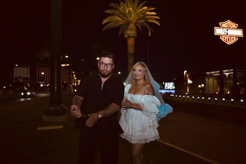 Couple walking at night on Las Vegas Strip near Harley-Davidson sign, woman in white dress with blue veil.