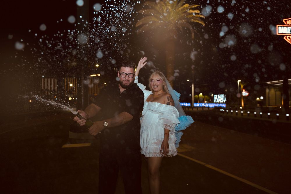 Couple celebrating on Las Vegas Strip at night with champagne spray, woman in white dress raising her hand.