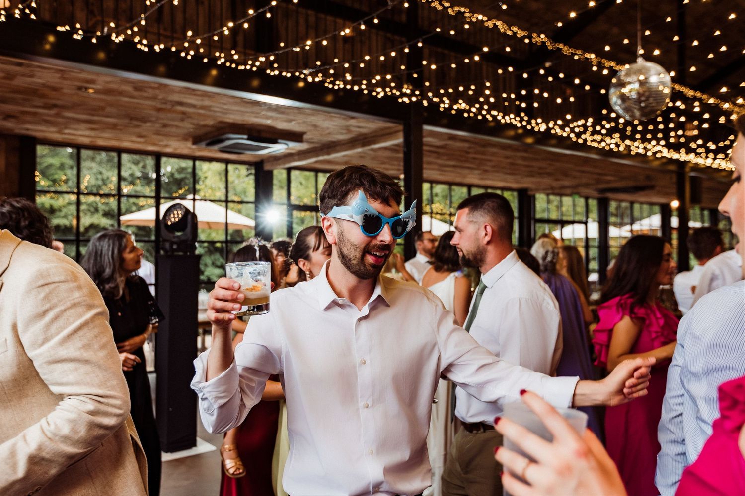 Two groomsmen in suits wearing sunglasses holding drinks outdoors at a wedding venue with white wisteria blooms.