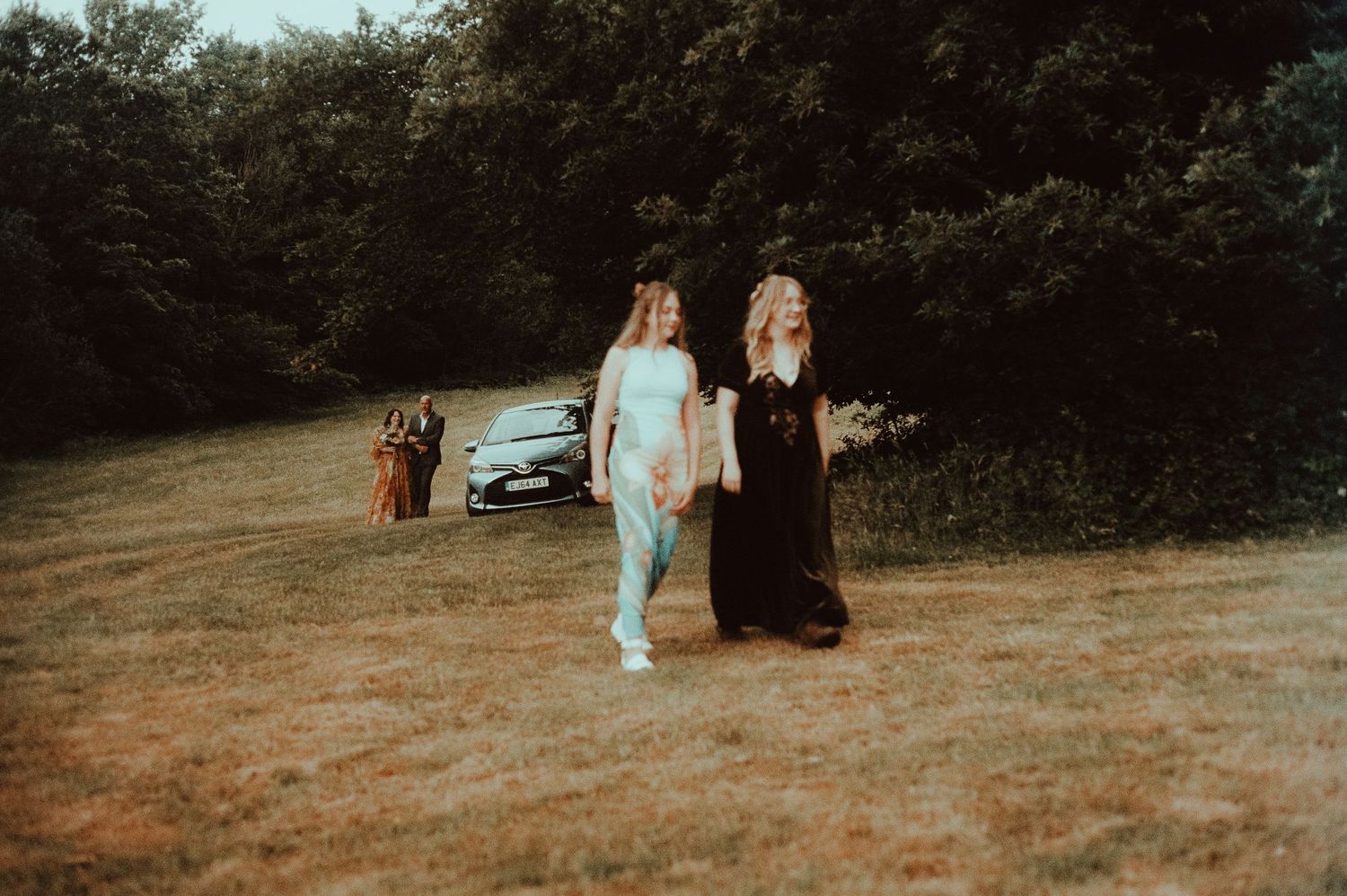 Two bridesmaids walking across a grassy field, one in light blue and one in black dress, with trees and car in background.