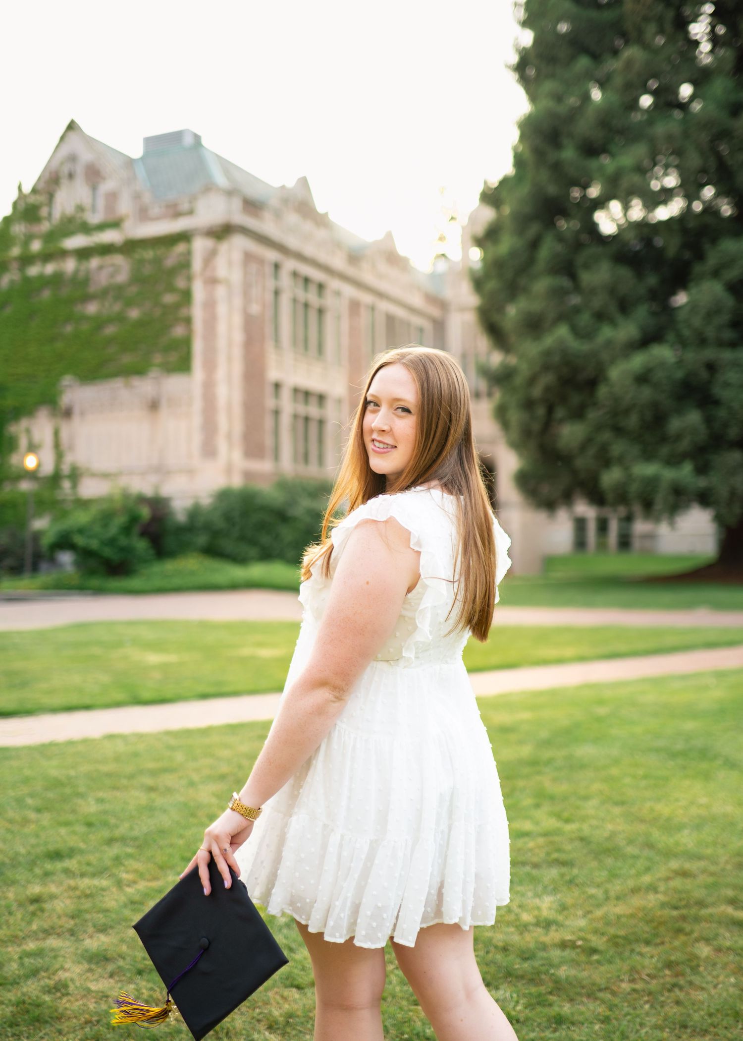 Smiling graduate in white dress holding cap on sunny campus lawn with historic brick building backdrop.