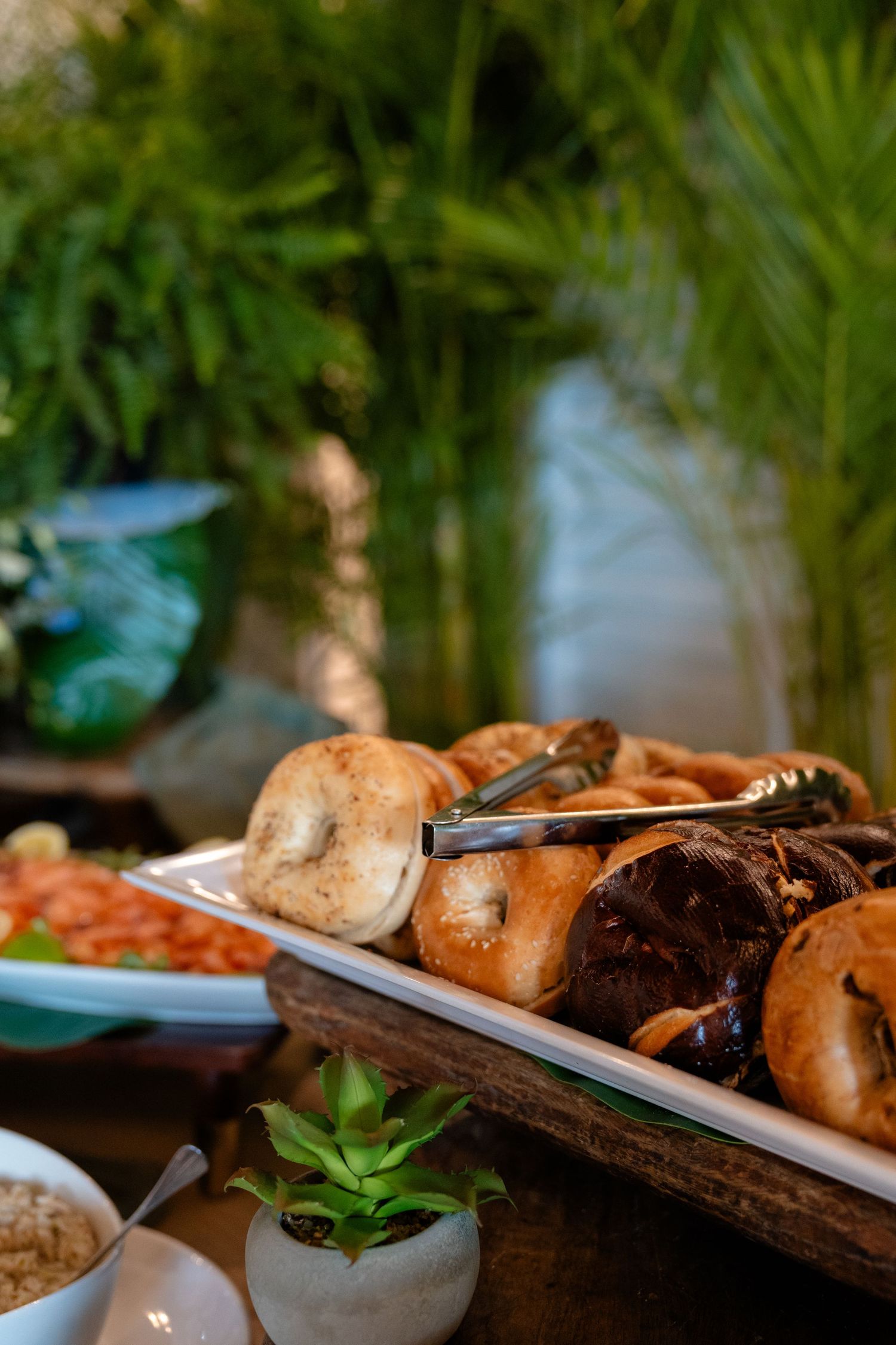 A buffet platter of assorted fresh mushrooms with serving tongs, surrounded by greenery and side dishes.