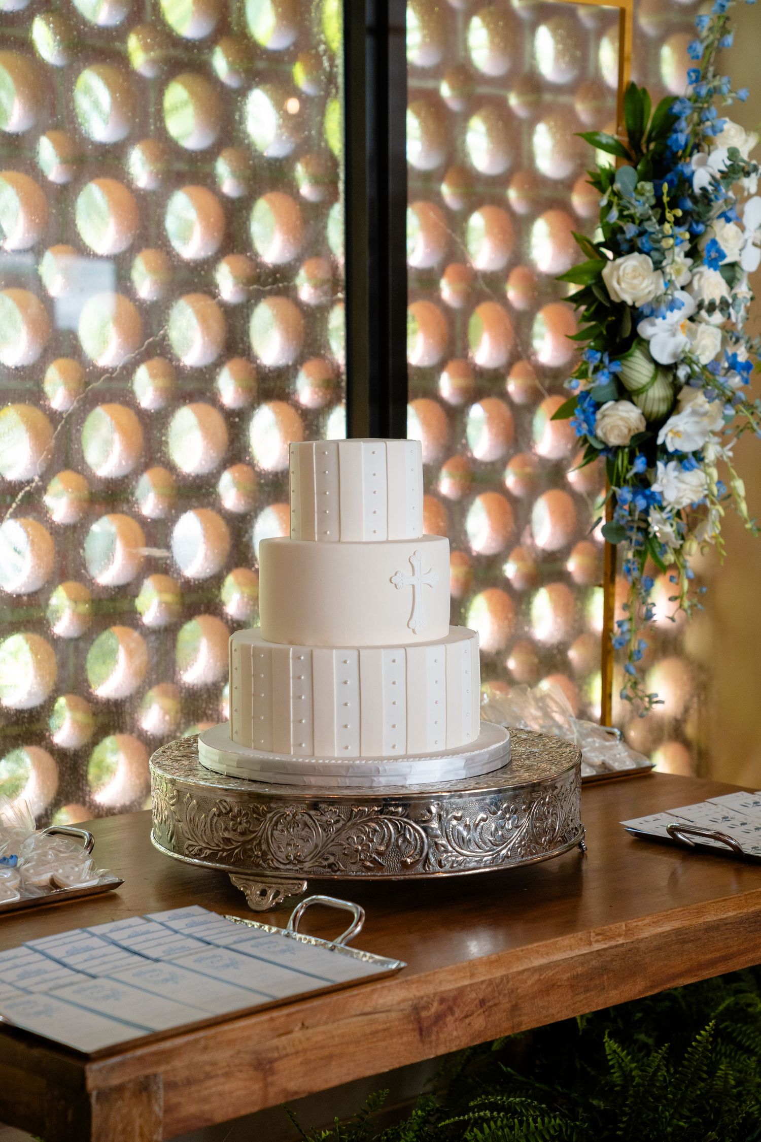 Elegant three-tier white wedding cake on silver stand, adorned with subtle details, displayed beside blue and white floral arrangement.