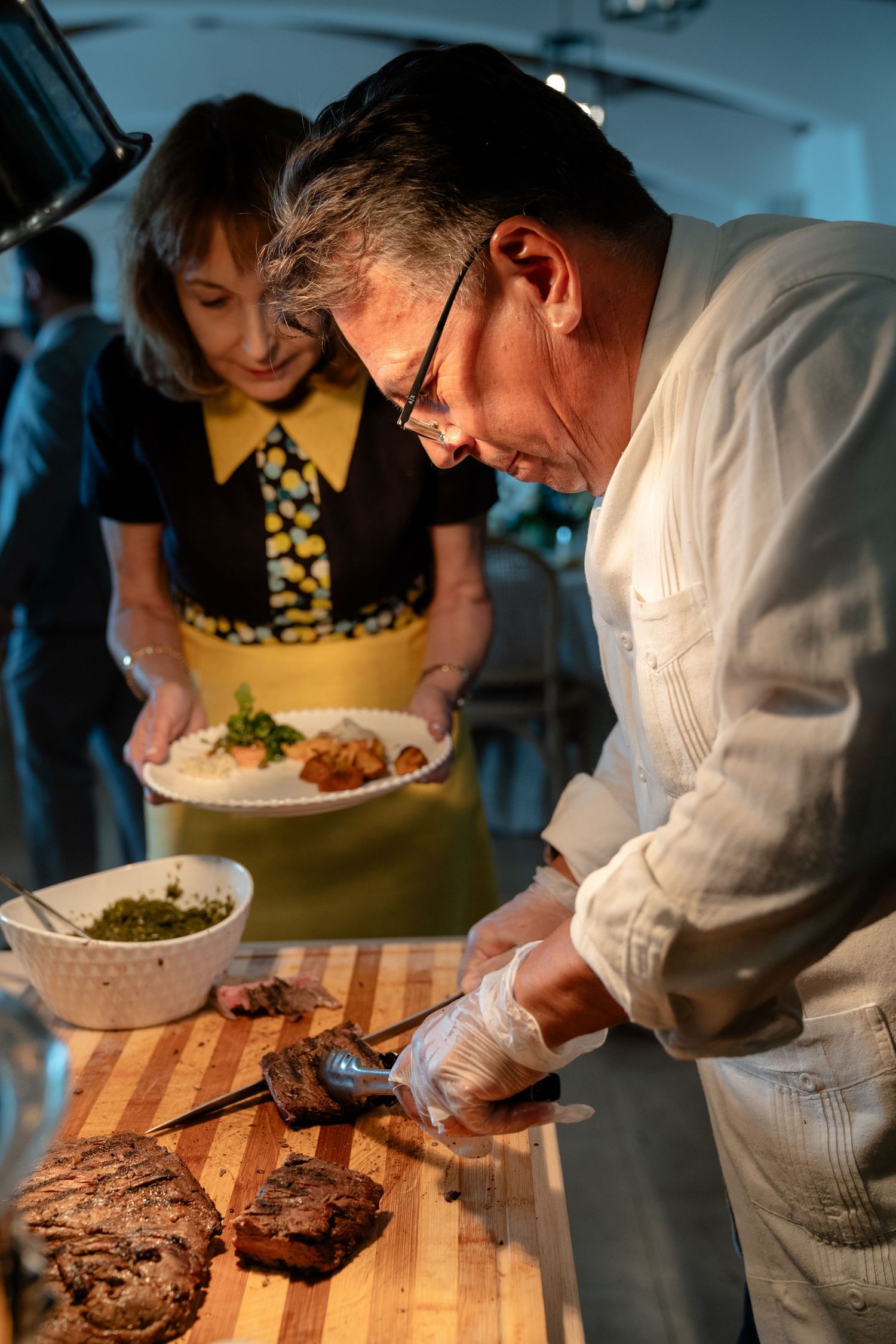 Chef carving grilled steak on wooden board while woman holds plate in professional kitchen setting.