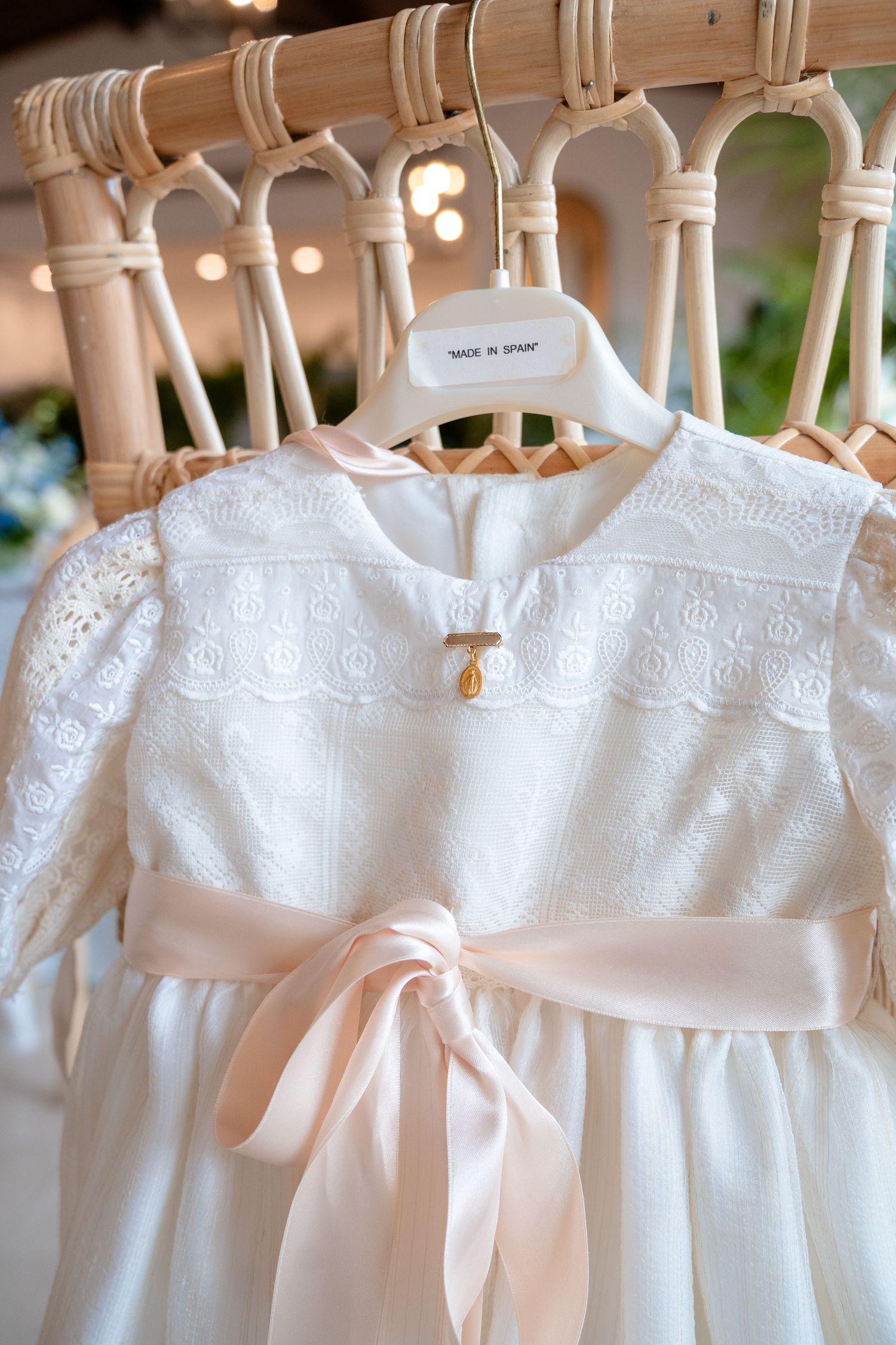 White lace baby dress with pink satin bow on wooden hanger, labeled Made in Spain, displayed on rattan rack.