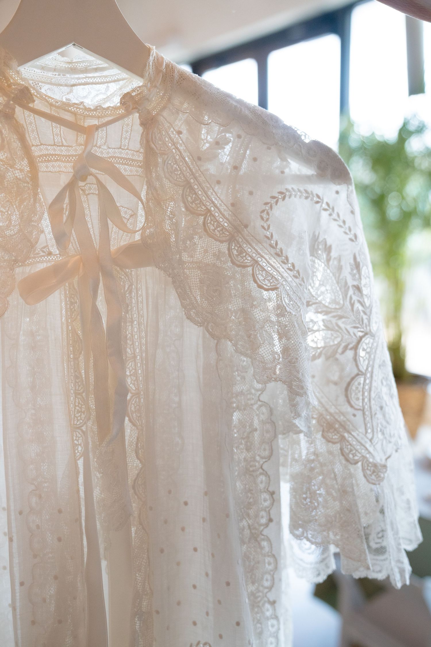 Delicate white lace bridal robe with floral embroidery and satin bow, hanging in soft natural light.