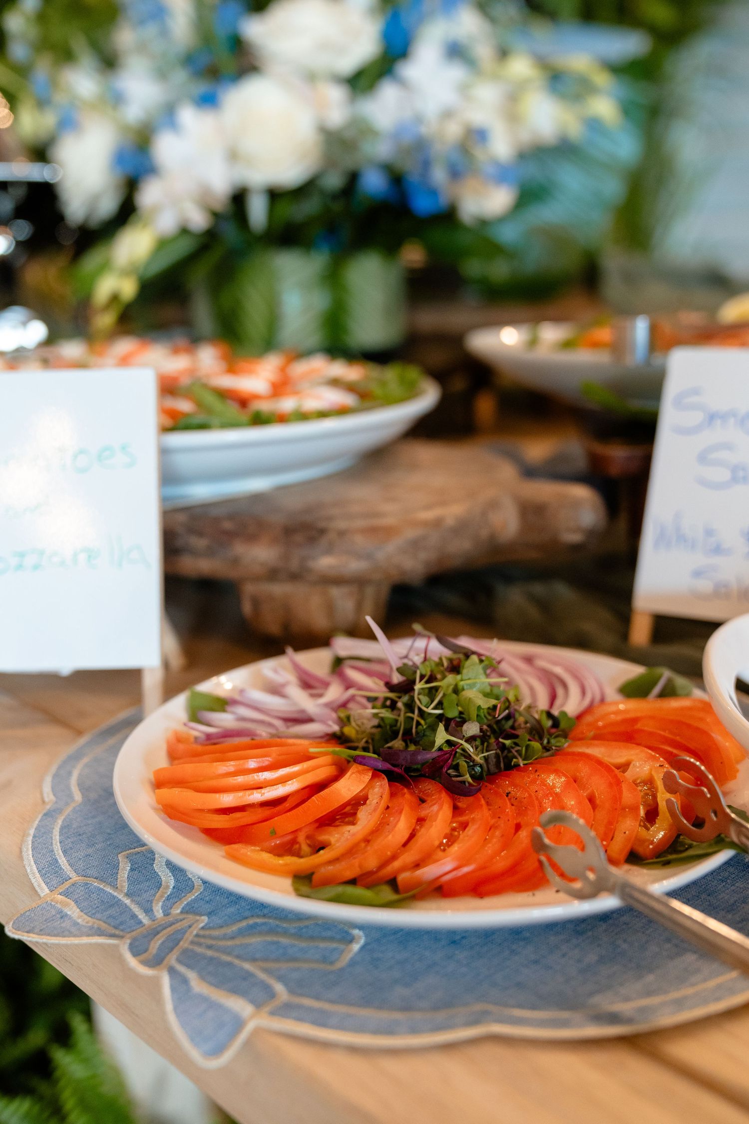 A colorful buffet plate with sliced tomatoes, red onions, and fresh greens on a decorative blue placemat at a catered event.
