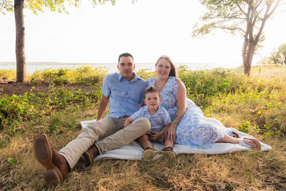 Family of three sitting on a blanket outdoors in golden hour light, smiling together near a scenic lakeside meadow.