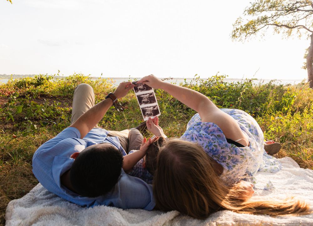 Couple lying on blanket outdoors holding ultrasound photos above them, sharing a tender pregnancy announcement moment.