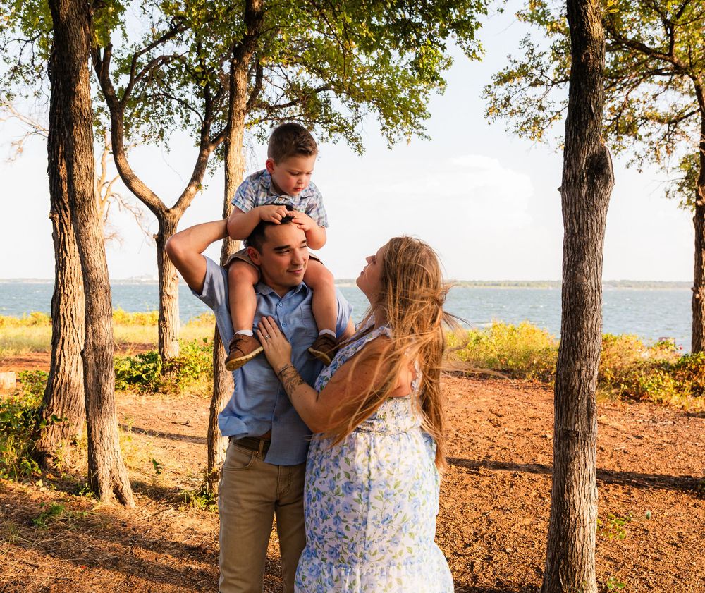 Family enjoying outdoor time near a lake, father carrying child on shoulders while mother embraces them among trees.