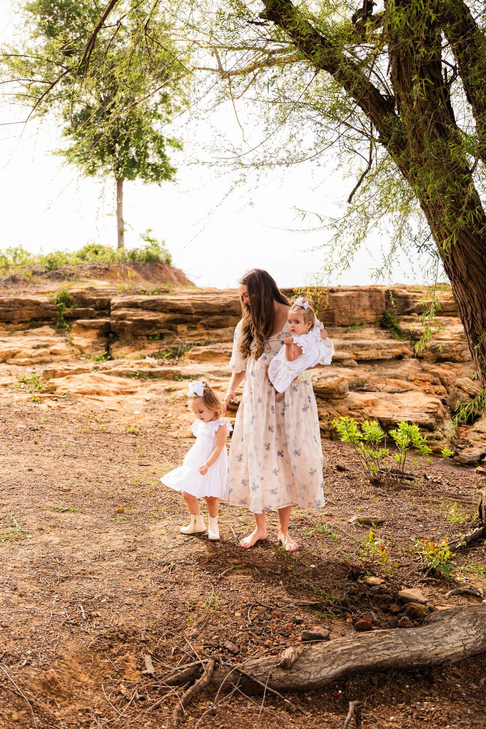 A mother in a floral dress holds a baby while a toddler in white stands beneath a tree near sandstone cliffs.