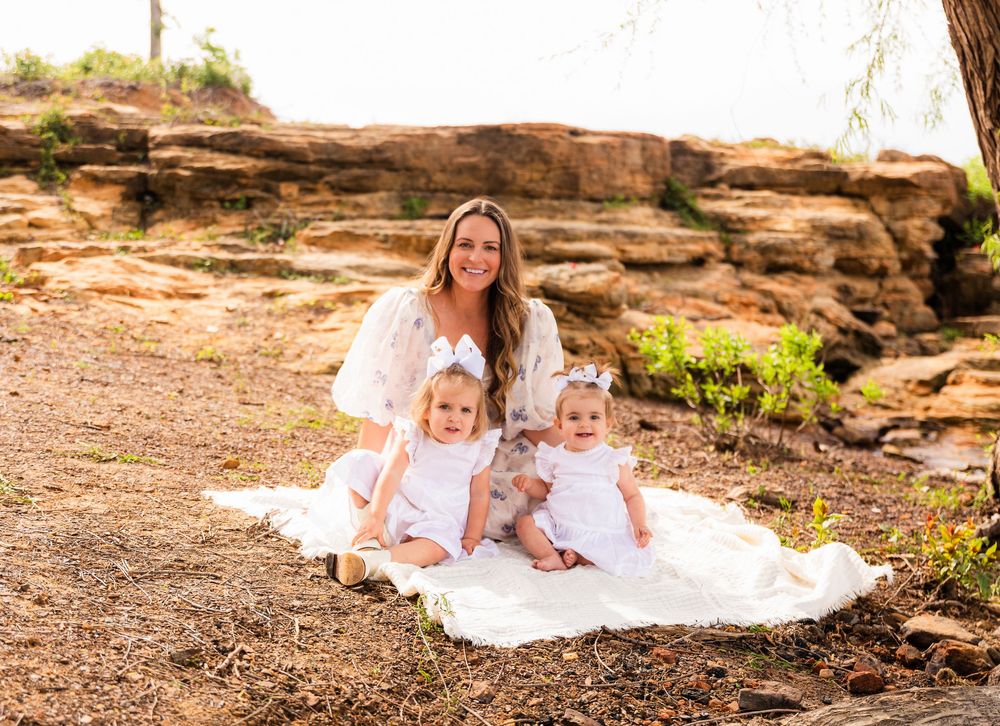 A woman in white dress sits on blanket with two toddlers near red rock formations outdoors.