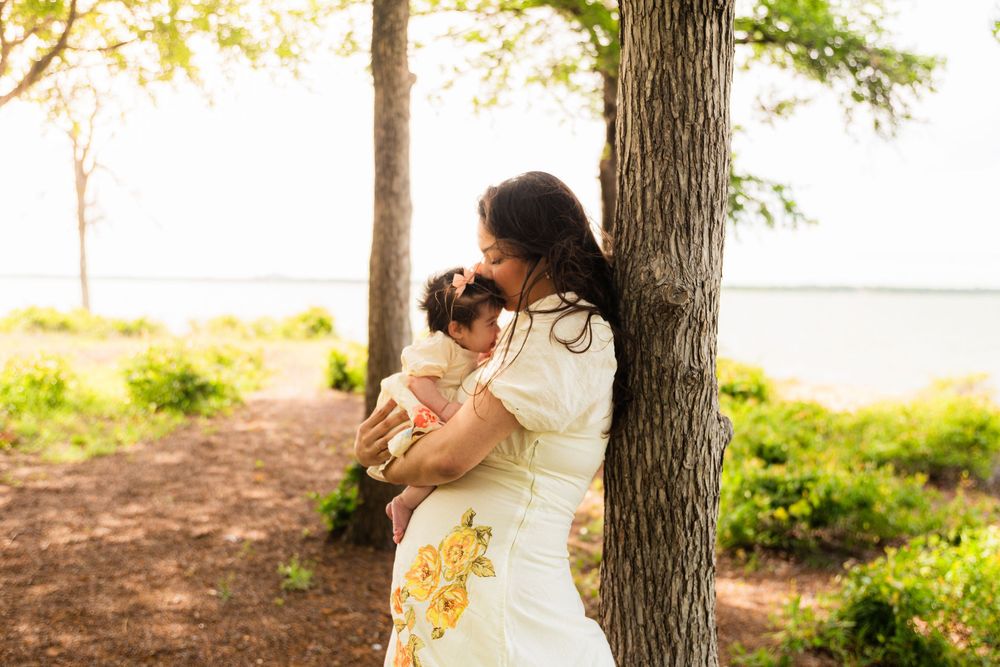 A mother in a floral dress tenderly holds her baby near a tree in a sunlit park by the water.