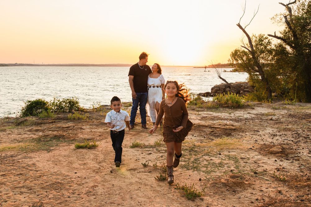 Family of four enjoying a golden sunset by the lake, with two children running on sandy shore.