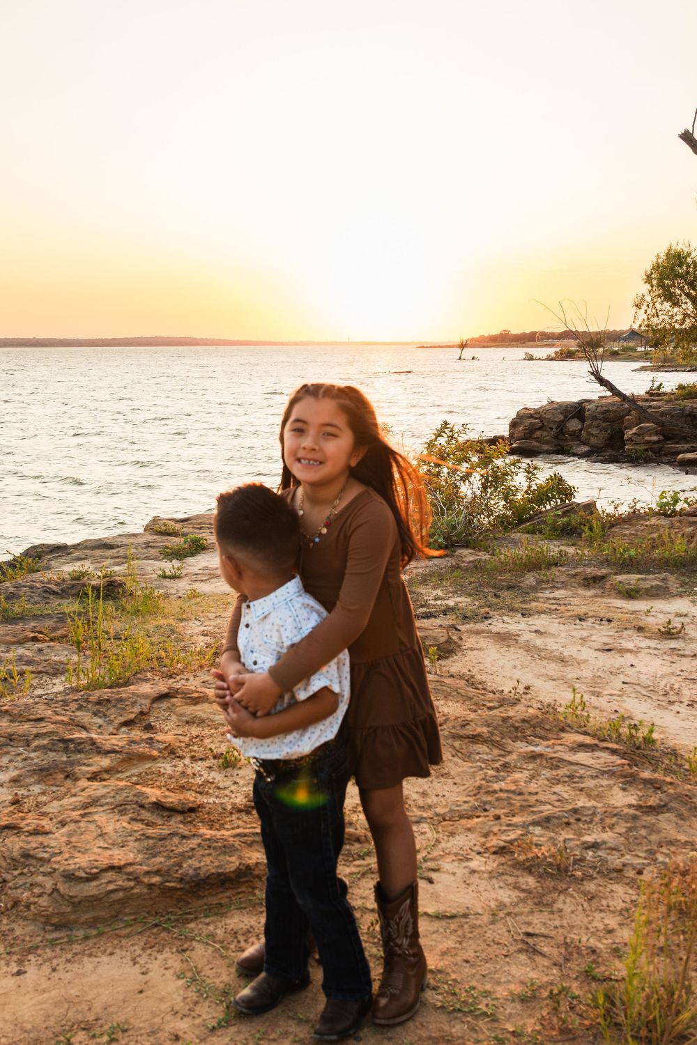 A girl hugs a young boy near a rocky lakeside at golden sunset, both smiling in casual outdoor attire.