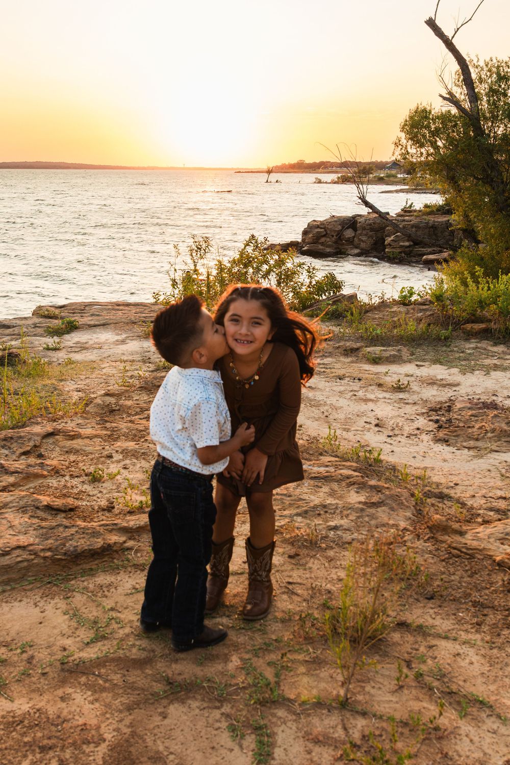 Two children share a sweet kiss at sunset by a rocky lakeside, with warm golden light reflecting on the water.