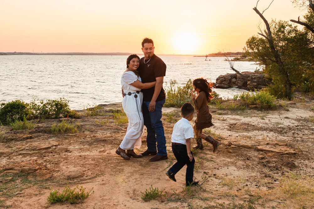 Family of four enjoying a golden sunset by the lake shore, parents embracing while children play nearby.